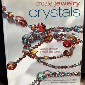 Könnte beinhalten: Ein Buchcover mit dem Titel "create jewelry crystals" in Rot und Blau. Das Cover zeigt farbenfrohen Perlenschmuck in Rot-, Orange- und Blautönen. Der Text lautet "Dazzling designs to make and wear" und die Autoren sind Marlene Blessing und Jamie Hogsett.