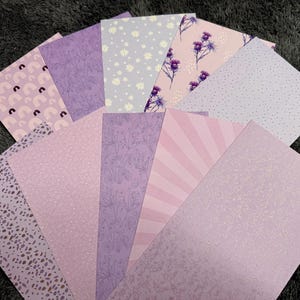 Könnte beinhalten: Eine Sammlung von Papieren mit Mustern in verschiedenen Lila- und Lavendeltönen. Designs umfassen Blumen-, Punkt-, Streifen- und abstrakte Muster. Die Blätter sind fächerförmig angeordnet und zeigen die Vielfalt der Designs.