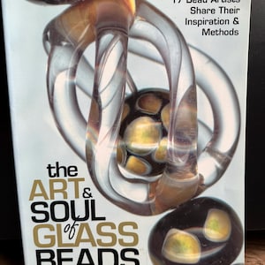 Könnte beinhalten: Ein Buch mit dem Titel "The Art & Soul of Glass Beads" mit einem Cover, das abstrakte Glasperlenkunst in Braun-, Gelb- und Klartönen zeigt. Der Text "17 Bead Artists Share Their Inspiration & Methods" ist ebenfalls sichtbar.