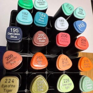 Puede incluir: Un conjunto de rotuladores de colores en un organizador negro. Los rotuladores tienen tapas triangulares con nombres y números de colores, como "Spectrum Green", "Teal Blue" y "Warm Fuchsia". Los rotuladores están dispuestos verticalmente.
