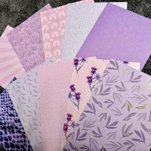 Könnte beinhalten: Eine Sammlung von Papieren mit Mustern in verschiedenen Lila- und Rosatönen. Designs umfassen Streifen, Blumenmuster, Punkte und ein Schlangenhautmuster. Einige Blätter weisen goldene Akzente auf. Ideal für Scrapbooking- oder Bastelprojekte.