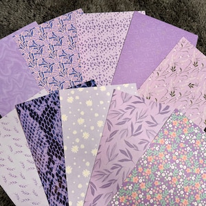 Könnte beinhalten: Eine Sammlung von Papieren mit verschiedenen Mustern in verschiedenen Lilatönen. Designs umfassen Blumen-, Blatt- und abstrakte Muster. Die Papiere sind fächerförmig angeordnet und zeigen die Vielfalt der Texturen und Stile.