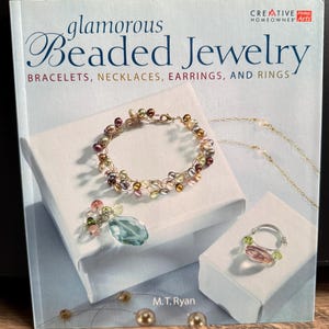 Könnte beinhalten: Ein Buch mit dem Titel "Glamorous Beaded Jewelry" mit Beispielen für Perlenarmbänder, Halsketten, Ohrringe und Ringe. Das Cover zeigt ein Perlenarmband, einen Anhänger mit einem blauen Stein und einen Ring. Das Buch ist von M.T. Ryan.