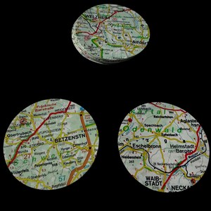 Op de afbeelding: Drie ronde objecten met kaartontwerpen, elk met een ander gedeelte van een gedetailleerde kaart. De kaarten bevatten wegen, steden en geografische details, met verschillende kleuren en tekst. Op een zwarte achtergrond.