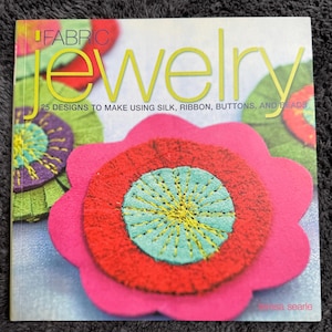Könnte beinhalten: Ein Buch mit dem Titel "Fabric Jewelry" mit einem floralen Design auf dem Cover. Das Cover zeigt eine große rosa Blume mit einem roten Zentrum und einem türkisfarbenen Zentrum. Der Titel des Buches ist in gelben und grünen Buchstaben.