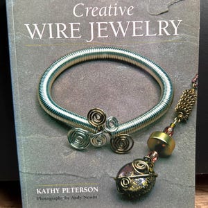 Könnte beinhalten: Buchcover "Creative Wire Jewelry" mit einem silbernen Drahtarmband mit dekorativen Drahtakzenten und einem Perlenanhänger. Das Buch ist von Kathy Peterson, Fotografie von Andy Newitt.