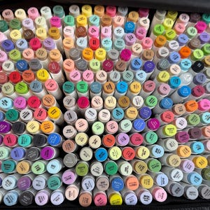 Puede incluir: Una gran colección de rotuladores de arte coloridos, cuidadosamente organizados en un estuche de transporte negro. Los rotuladores tienen barriles blancos y tapas en varios tonos, incluyendo rosa, azul, verde, amarillo y morado. La imagen muestra una amplia gama de colores.