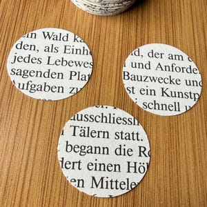 Op de afbeelding: Drie witte, cirkelvormige papieren uitsparingen met tekst uit een boek liggen op een houten oppervlak. De tekst is in het Duits. Een stapel vergelijkbare uitsparingen is zichtbaar op de achtergrond. Ideaal voor knutselen of scrapbooking.