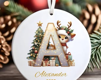 Puede incluir: Adorno navideño redondo y blanco con un diseño festivo. Presenta la letra "A" con un árbol de Navidad y un reno con sombrero. El nombre "Alexander" y el año "2025" están escritos en oro.