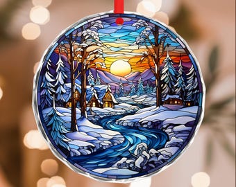 May include: Round Christmas ornament with a winter landscape scene. The design features a river, snow-covered trees, and cabins, with a sunset in the background. The ornament is edged with a silver border and has a red ribbon for hanging.