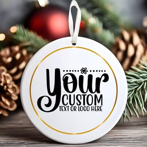 May include: White round Christmas ornament with gold trim and a white ribbon. The ornament has the words "Your Custom Text or Logo Here" in black script and block letters. The background includes pine needles, pine cones, and a red ornament.