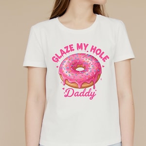 May include: White t-shirt featuring a pink frosted donut graphic with sprinkles. The text "GLAZE MY HOLE" arches above the donut, and "Daddy" is written below in pink script. The shirt has a crew neck and short sleeves.