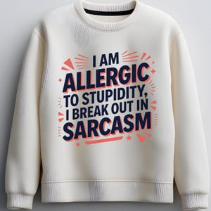 May include: Cream-colored sweatshirt with the text "I AM ALLERGIC TO STUPIDITY, I BREAK OUT IN SARCASM" in navy blue and coral lettering. The design includes star and triangle accents.