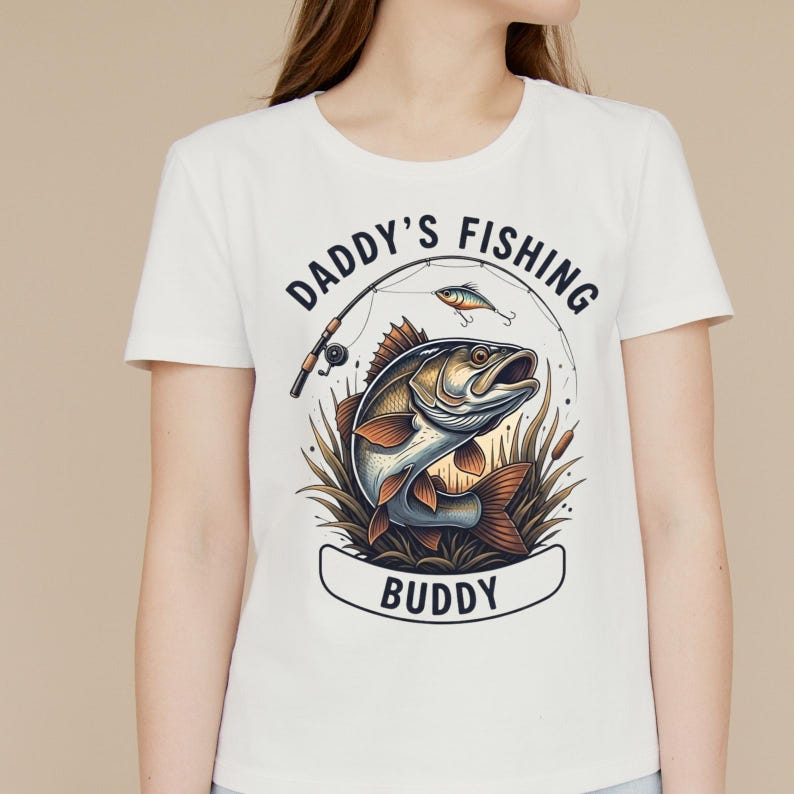 K&ouml;nnte beinhalten: Wei&szlig;es T-Shirt mit einem Angelmotiv. Das Design zeigt einen Fisch, der aus dem Wasser springt, eine Angelrute und einen K&ouml;der. Der Text "DADDY'S FISHING BUDDY" ist &uuml;ber dem Fisch zu sehen. Die Grafik verwendet Braun-, Blau- und Gr&uuml;nt&ouml;ne.