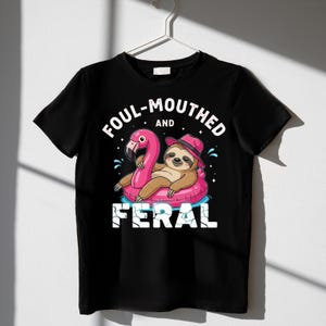 May include: Black t-shirt with a graphic of a sloth wearing a pink hat and floating on a pink flamingo pool float. The text on the shirt reads "FOUL-MOUTHED and FERAL".