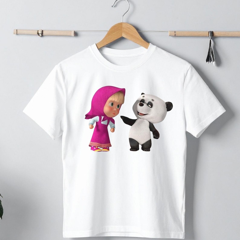 K&ouml;nnte beinhalten: Wei&szlig;es T-Shirt mit einer Cartoon-Illustration eines M&auml;dchens in einem rosa Kleid und einem Panda. Das M&auml;dchen tr&auml;gt eine rosa Kapuze und ein Kleid. Der Panda ist schwarz und wei&szlig;. Das Design ist auf dem Shirt zentriert.