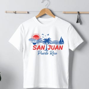 以下が含まれることがあります： 白いTシャツにカラフルなグラフィックデザイン。デザインには、赤い太陽、青い波、ヤシの木、ヨット、そして「SAN JUAN Puerto Rico」の文字が赤と青で描かれています。Tシャツは木製のハンガーに掛けられています。
