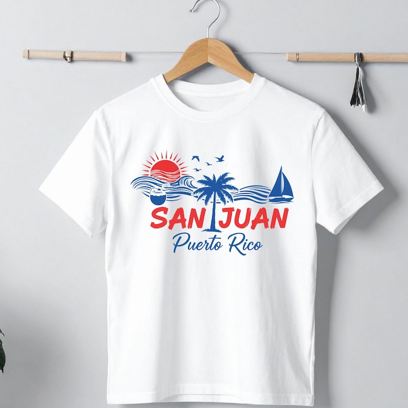 以下が含まれることがあります： 白いTシャツにカラフルなグラフィックデザイン。デザインには、赤い太陽、青い波、ヤシの木、ヨット、そして「SAN JUAN Puerto Rico」の文字が赤と青で描かれています。Tシャツは木製のハンガーに掛けられています。