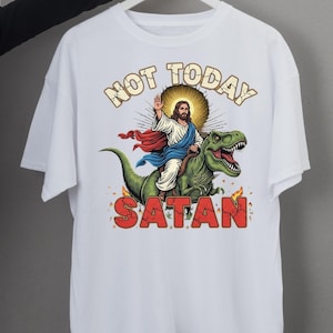 May include: White t-shirt featuring a graphic of Jesus riding a green dinosaur, with the words "NOT TODAY SATAN" in a bold font. Jesus is depicted with a halo, wearing a white robe and red and blue cape. Flames are at the bottom of the graphic.