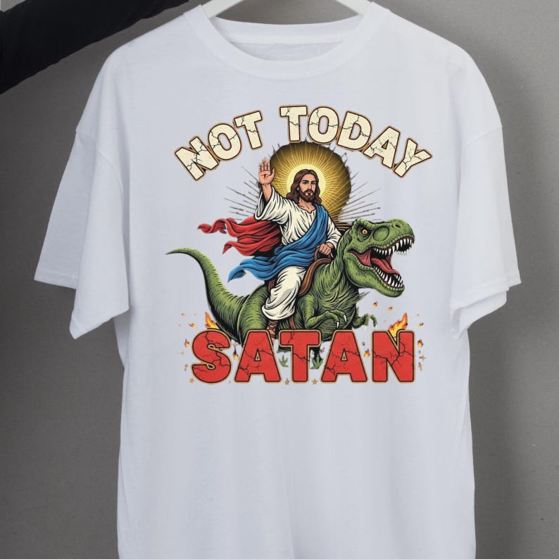 May include: White t-shirt featuring a graphic of Jesus riding a green dinosaur, with the words "NOT TODAY SATAN" in a bold font. Jesus is depicted with a halo, wearing a white robe and red and blue cape. Flames are at the bottom of the graphic.