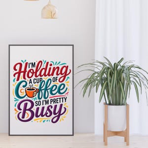 May include: A framed art print with the words "I'm holding a cup of coffee so I'm pretty busy" in colorful lettering. The print is in a black frame and is displayed next to a potted plant.