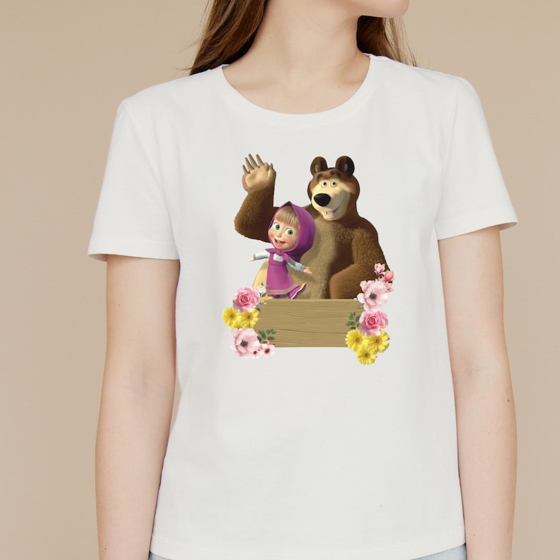 K&ouml;nnte beinhalten: Wei&szlig;es T-Shirt mit einer Cartoon-Illustration eines B&auml;ren und eines M&auml;dchens, mit einem Holzschild und Blumendekorationen. Der B&auml;r ist braun und das M&auml;dchen tr&auml;gt ein rosa Kleid. Das Design ist mittig platziert.
