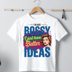 May include: White t-shirt with the text "I'M NOT BOSSY I just have Better IDEAS" in blue, green, and purple. A graphic of a woman with brown hair is on the right side of the shirt. The shirt is hanging on a wooden hanger.