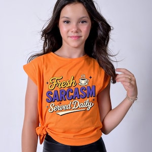 May include: Orange t-shirt with the text "Fresh Sarcasm Served Daily" in yellow and purple. The shirt has a coffee cup graphic and is knotted at the waist. The model is wearing black pants.