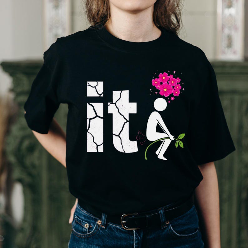 May include: Black t-shirt with a white graphic that reads "it" in a cracked font. A stick figure is depicted squatting and using leaves as a spray, with pink flowers above. The shirt is worn by a person.