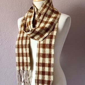 May include: A brown and cream-colored plaid scarf with fringe. The scarf is displayed on a mannequin. The scarf has a woven texture and a classic checkered pattern. The fringe hangs at the bottom of the scarf.