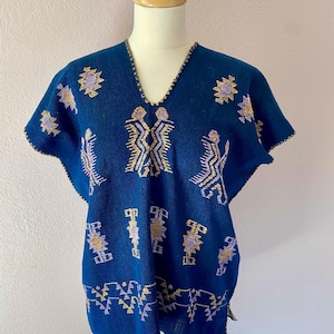 Op de afbeelding: Een donkerblauwe tuniek met korte mouwen en een V-hals. De tuniek is versierd met geborduurde geometrische en gestileerde menselijke figuren in goud en lavendel. De randen zijn afgezet met goudkleurig garen. Het kledingstuk wordt op een mannequin gepresenteerd.