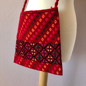 May include: Red embroidered crossbody bag with a geometric pattern in black, pink, and green. The bag has a long strap and a flap closure.