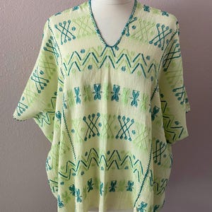 May include: A light yellow poncho featuring a V-neckline and short sleeves. The garment is decorated with an intricate embroidered pattern in shades of teal and lime green, consisting of horizontal zigzag lines and repeating geometric motifs.