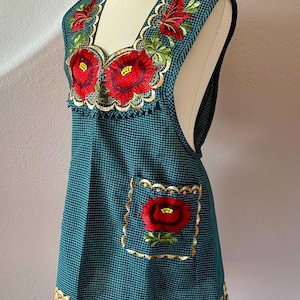 May include: Teal apron with a grid pattern, featuring embroidered red flowers with green leaves and gold trim. The apron has a pocket with a flower and a scalloped neckline and hem. The apron is displayed on a mannequin.
