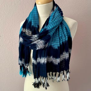 May include: A blue and white patterned scarf with fringe. The scarf is draped over a mannequin. The scarf has a mix of blue shades, including turquoise and navy, with white accents. The fringe at the ends is a mix of white and blue.