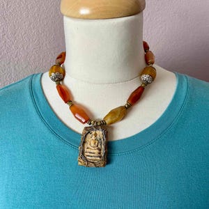 Puede incluir: Un collar hecho de cuentas de cornalina con un colgante de piedra tallada que representa una imagen de Buda. Las cuentas son de una mezcla de colores naranja y marrón, y el colgante es de color marrón claro.