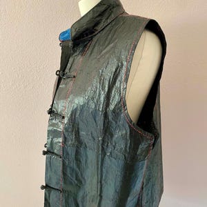 Puede incluir: Un chaleco negro sin mangas con cuello mandarín y cierres de rana. El chaleco tiene costuras rojas y un aspecto texturizado. La prenda se exhibe en un maniquí sobre un fondo rosa claro. El chaleco parece estar hecho de un material arrugado.