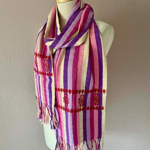 May include: A colorful scarf with vertical stripes in shades of pink, purple, and cream, displayed on a mannequin. The scarf features embroidered red heart-shaped patterns and fringe at the ends. A stylish accessory.