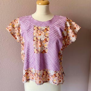 May include: A lavender blouse with a floral print and a geometric pattern. The blouse has short sleeves and a round neckline.