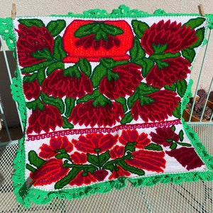 May include: A white textile with a vibrant floral design in red, green, and orange. The flowers are intricately embroidered, and the edges are trimmed with a green crocheted border. The textile is hanging on a light blue metal chair.
