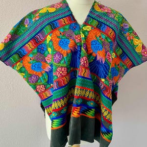 May include: A vibrant, hand-embroidered poncho with a V-neck. The textile features a colorful design with birds, flowers, and geometric patterns in shades of blue, orange, green, and red. The bottom hem is a dark gray.