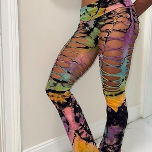 Op de afbeelding: Tie-dye legging in een mix van paars, geel, groen en zwart. De legging heeft een uniek ontwerp met uitgesneden delen die bij elkaar worden gehouden door metalen ogen, wat een opvallende look creëert. De stof lijkt zacht en rekbaar.