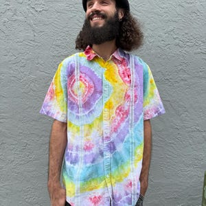 May include: A short-sleeved button-down shirt featuring a vibrant tie-dye pattern in shades of yellow, pink, purple, and light blue. The shirt includes two vertical white lace panels running down the front, a standard collar, and a button-up front.