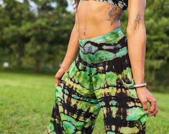 May include: A two-piece outfit featuring a tie-dye pattern in shades of green, black, and yellow. The top is a crop top with thin straps, and the pants are loose-fitting with a gathered waistband. The outfit is worn outdoors.