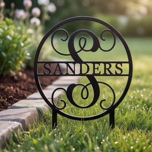 May include: A black circular metal garden sign featuring a large decorative letter S in the center. A horizontal bar runs across the middle of the circle with the name SANDERS in capital letters. The sign has two stakes at the bottom base.