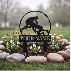 May include: A black metal garden stake featuring a circular frame with a silhouette of two wrestlers in a grappling position. Below the circular frame is a rectangular plaque with the text YOUR NAME in bold, capitalized, light-colored letters.
