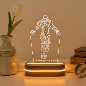 May include: A decorative Iron Man-themed LED lamp. The lamp features a white outline of the superhero with the text "I love you 3000" on the base. The lamp is on a wooden base with a warm light.