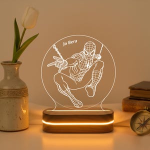 May include: A Spiderman-themed LED lamp with a circular design, featuring the superhero in a dynamic pose. The lamp has a wooden base with a warm, glowing light. The text "Jo Bera" is visible on the lamp.