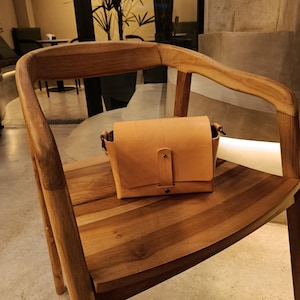 May include: A small, tan leather crossbody bag with a flap closure and a strap, resting on a wooden chair. The bag has a rectangular shape and a simple, classic design. The chair is made of light-colored wood.