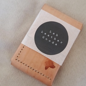 May include: A light brown leather wallet with a stitched edge and a dog silhouette. The wallet is wrapped with a white paper band and a black circular label that reads "Lab Leather Goods".